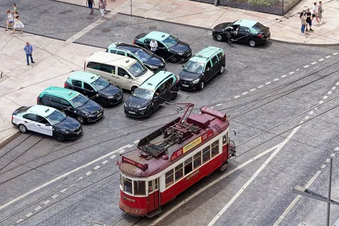 Ett tiotal beige och svarta taxibilar står uppradade på ett torg, sett snett uppifrån, medan en röd spårvagn passerar