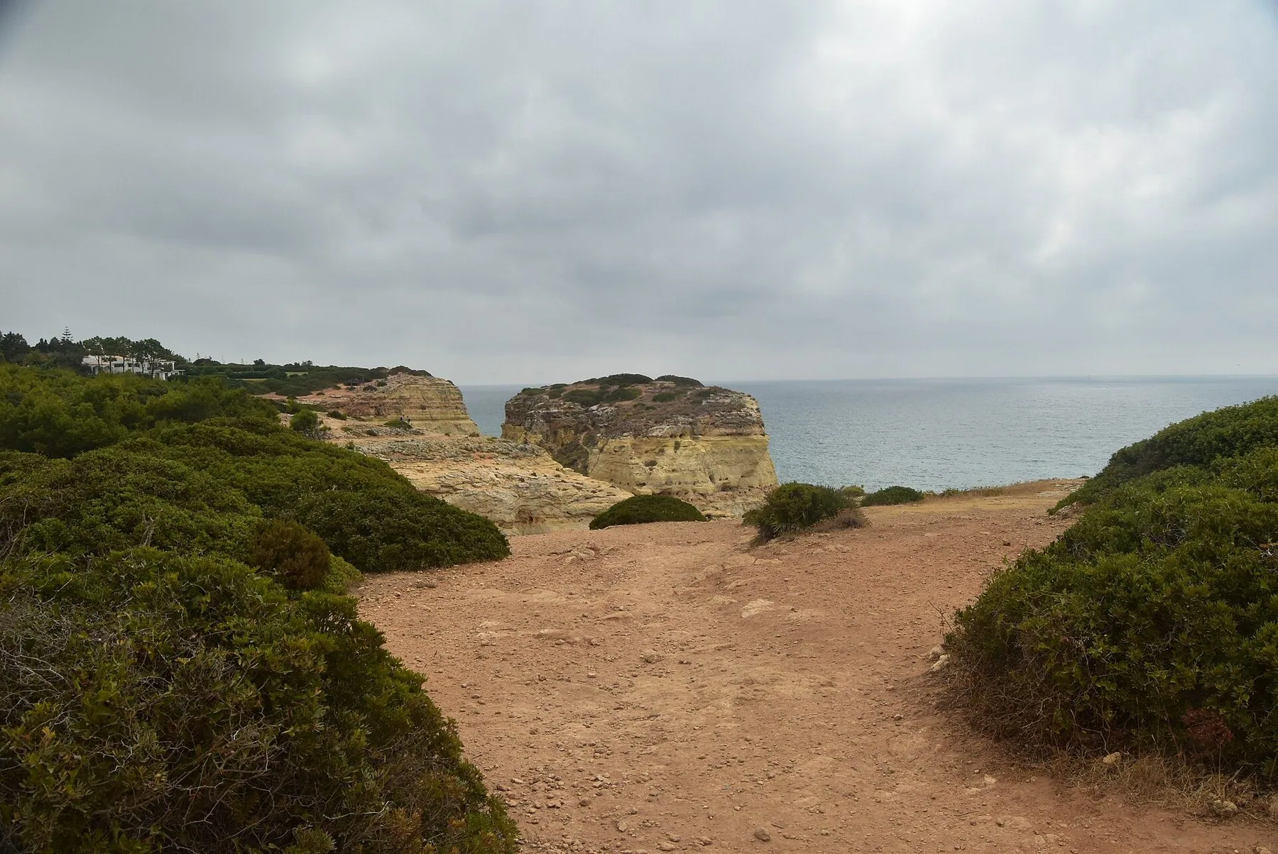 Den röda grusstigen på leden Sete Vales Suspensos går genom låg buskvegetation fram mot klippkanten, med en flat kalkstensudde och havet framför.